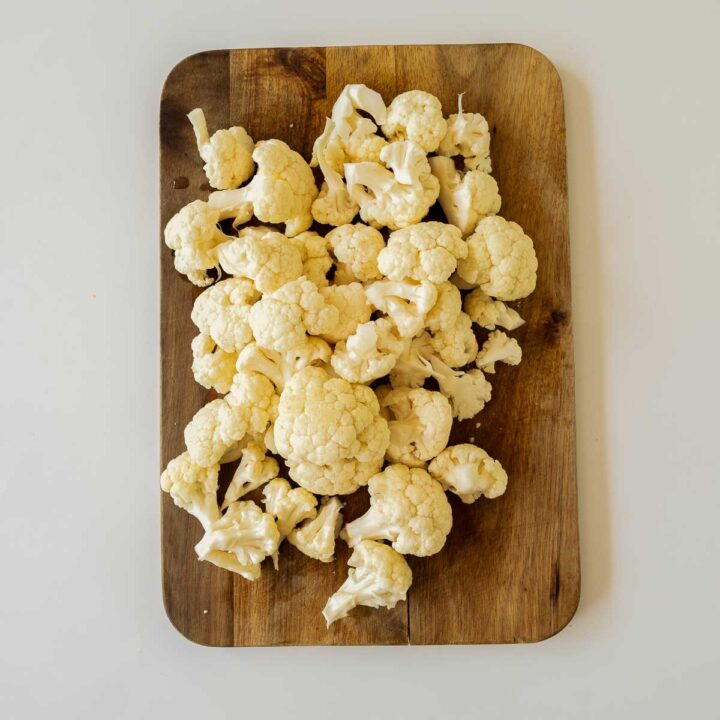 Overhead view of a pile of raw, bite-sized cauliflower florets scattered across a rustic rectangular wooden cutting board.