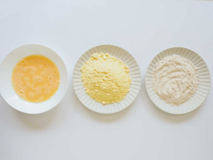 Overhead view of three white plates laid out side-by-side for a standard breading process: the first plate holds beaten eggs (egg wash), the middle plate holds a pile of yellow flour (cornmeal or chickpea flour), and the third plate holds a pile of light brown flour (all-purpose or whole wheat flour).