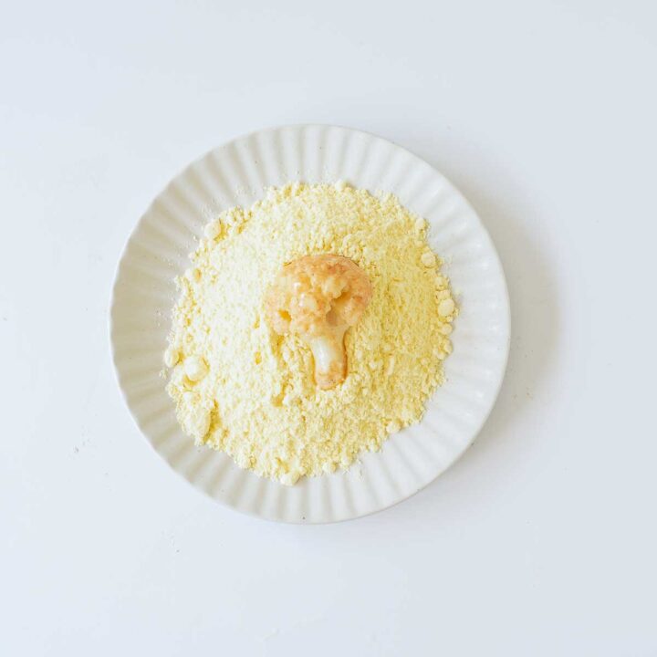 Overhead close-up of a white fluted plate covered in yellow flour (likely cornmeal or chickpea flour), with a single cauliflower floret that has been dipped in liquid (egg wash) resting in the center, ready to be coated.