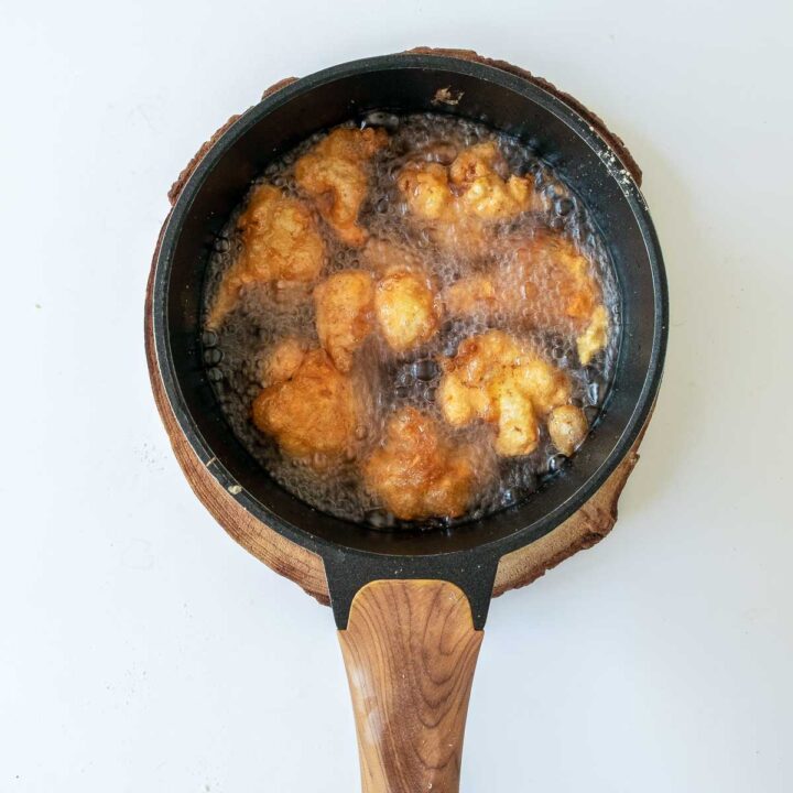 Overhead shot of several battered cauliflower florets being deep-fried in bubbling hot oil inside a black skillet with a wooden handle, turning golden brown.