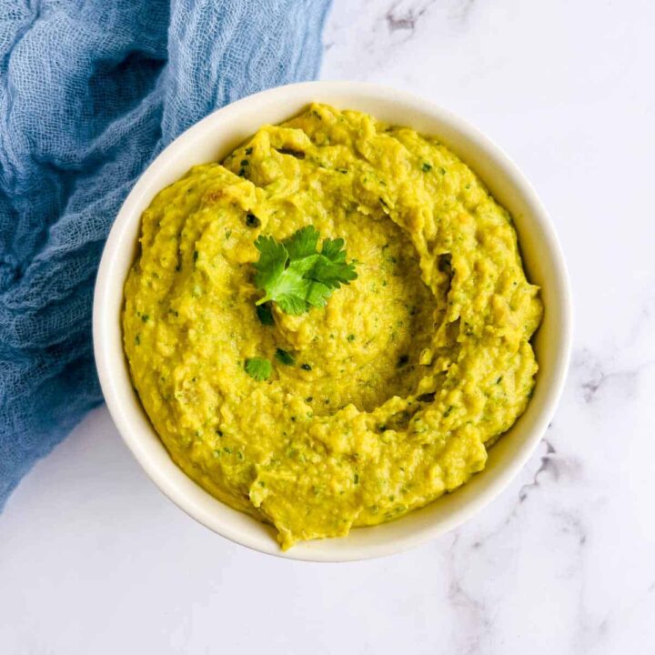 An overhead shot of a bowl of thick, bright yellow curried bean dip, garnished with a sprig of cilantro, sitting on a white marble surface next to a crumpled blue cloth napkin.