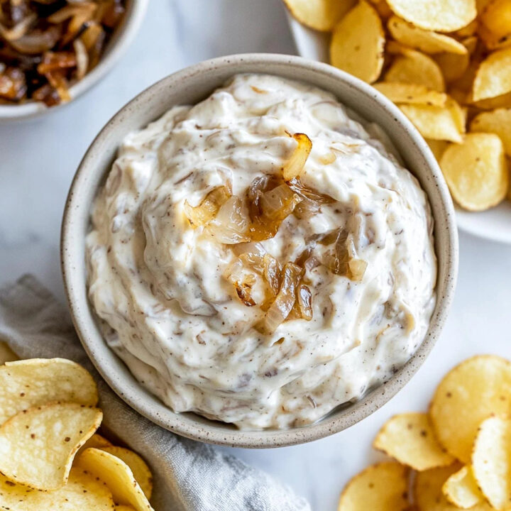 An overhead close-up shot of a bowl of creamy French onion Greek yogurt dip, topped with a small spoonful of caramelized onions. The dip is surrounded by potato chips and a white bowl of additional caramelized onions in the background.