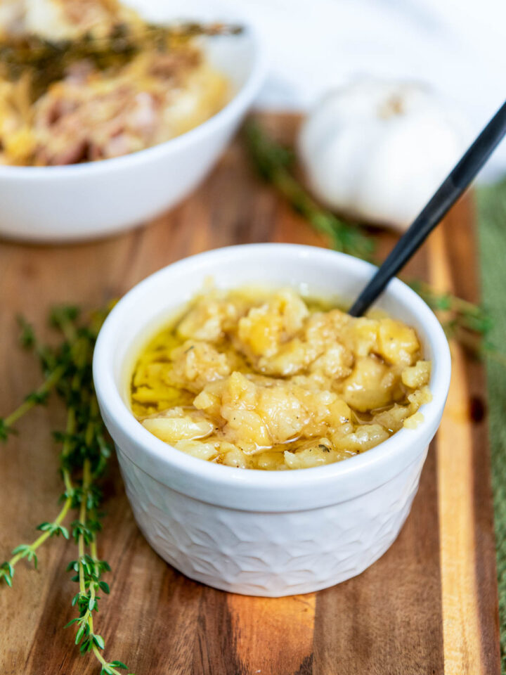 A white ramekin filled with soft, golden garlic confit submerged in olive oil, served on a wooden board with fresh thyme and a bulb of raw garlic.