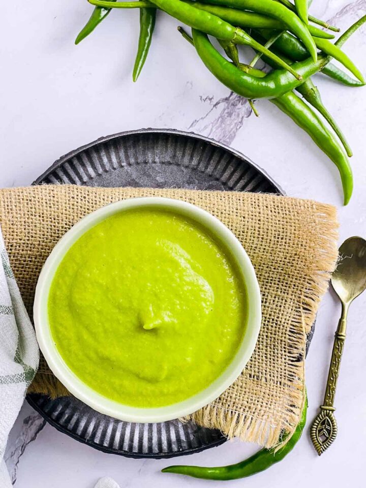 A small white bowl of smooth, bright green chili sauce or chutney, placed on a dark, textured plate with a gold spoon and fresh green chili peppers.