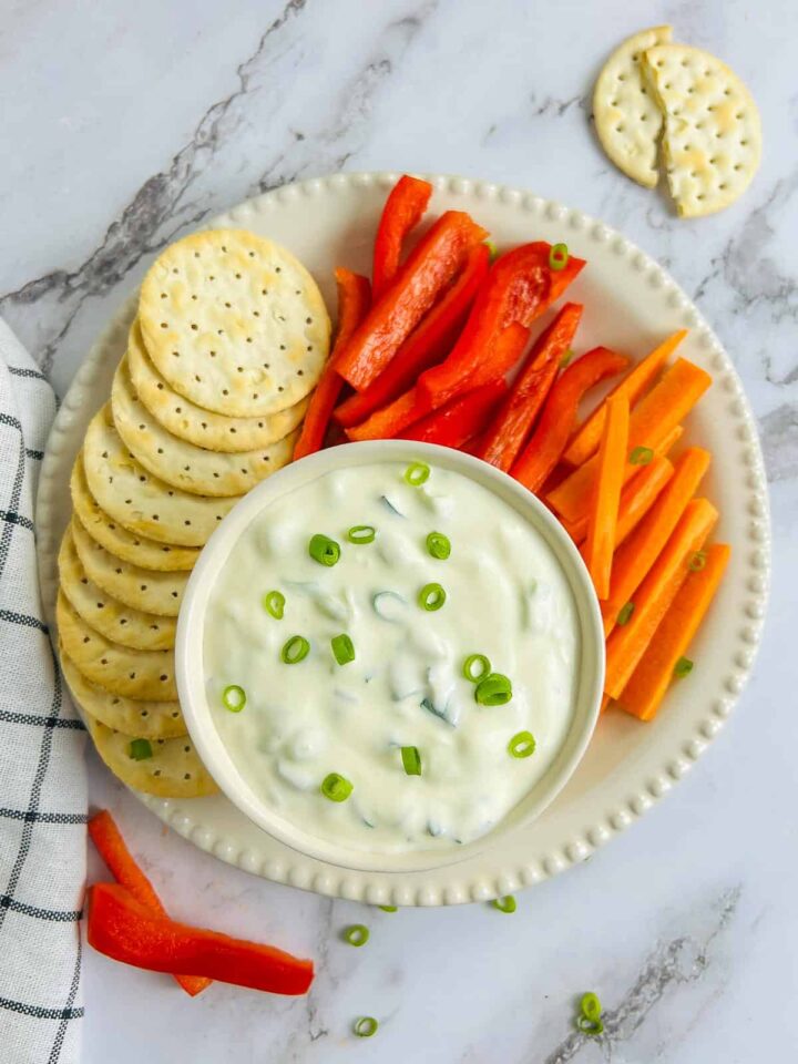 An overhead shot of a creamy white green onion dip in a small bowl, garnished with sliced green onions. The bowl sits on a white platter surrounded by a stack of round crackers, red bell pepper strips, and carrot sticks.