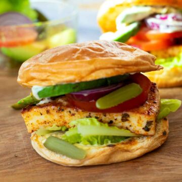 A close-up of a halloumi cheese burger on a toasted bun, layered with lettuce, tomato, red onion, cucumber, and pickles.