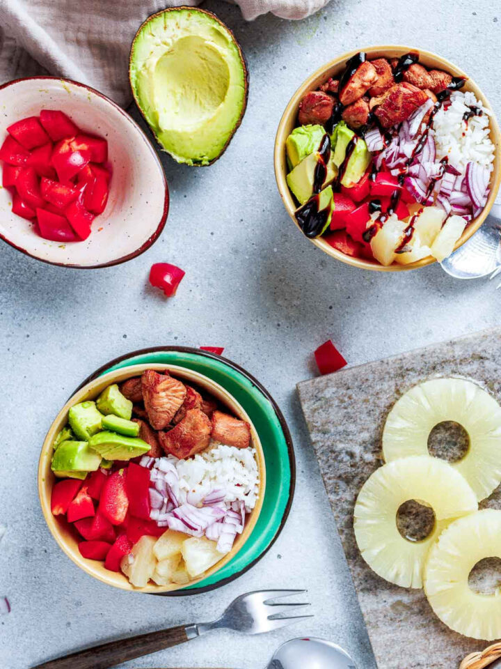 Overhead view of two colorful Hawaiian Chicken Bowls and surrounding fresh ingredients, including: white rice, cooked chicken, diced red bell pepper, red onion, pineapple, and avocado. One bowl is drizzled with a dark glaze.
