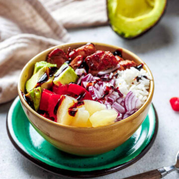 Close-up of a single Hawaiian Chicken Bowl filled with white rice, dark-glazed chicken pieces, diced red onion, red bell pepper, cubed pineapple, and avocado, all drizzled with a dark sauce and served in a gold bowl on a vibrant green saucer.