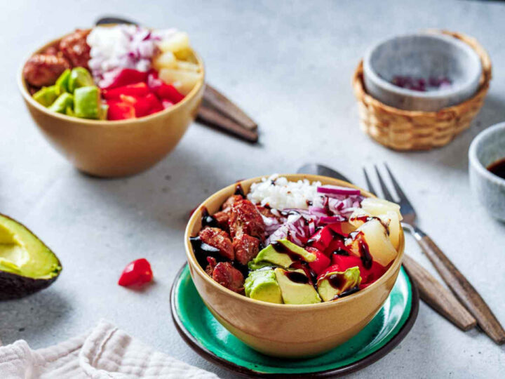 Two bowls of Hawaiian Chicken Bowl, with the one in the foreground showing white rice, chicken, avocado, red onion, red pepper, and pineapple drizzled with a dark sauce, set on a green saucer with a fork and spoon nearby.