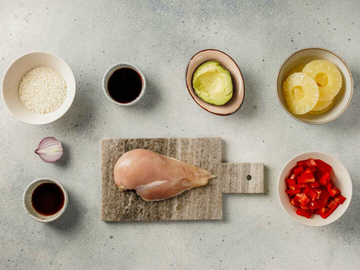 Overhead flat lay of ingredients for a Hawaiian Chicken Bowl, arranged on a light gray surface: a raw chicken breast sits on a stone cutting board, surrounded by bowls of uncooked white rice, sliced pineapple rings, diced red bell pepper, a halved avocado, a sliced red onion, and two small bowls containing a dark liquid (likely soy sauce, teriyaki, or balsamic glaze).
