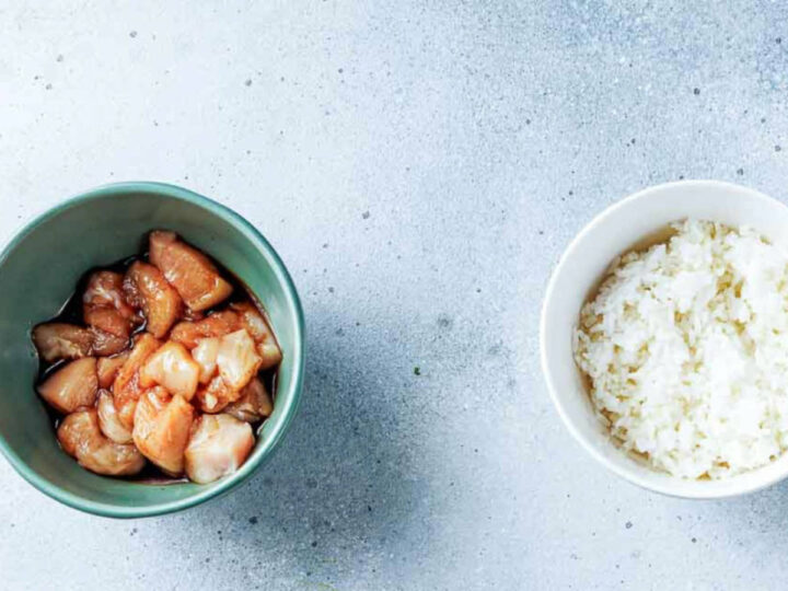 An overhead shot showing two bowls on a light gray surface: a teal bowl on the left holds cubed chicken pieces marinating in a dark sauce (likely soy or teriyaki), and a white bowl on the right holds cooked white rice.