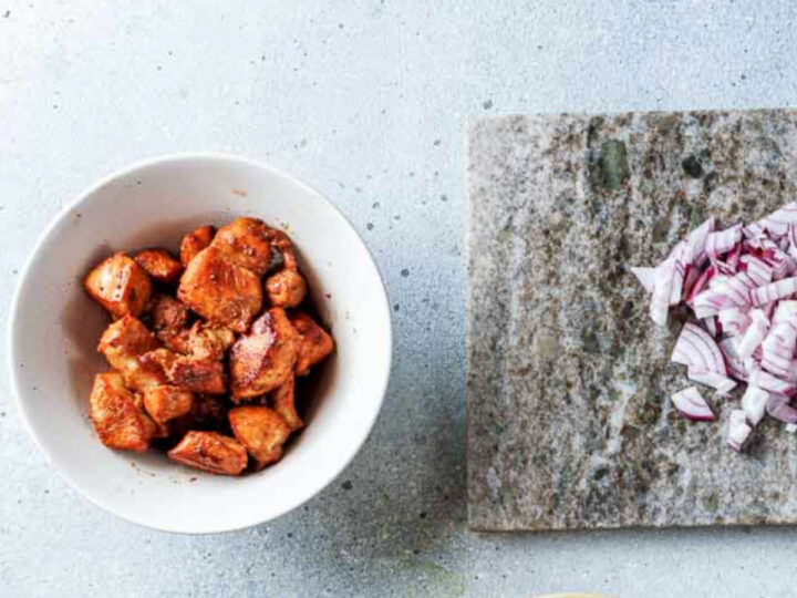 An overhead shot showing a white bowl of cooked, browned, and seasoned cubed chicken next to a gray stone cutting board with a pile of chopped red onion.