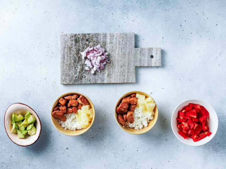 Overhead shot showing the assembly process for two Hawaiian Chicken Bowls. A stone cutting board with diced red onion is at the top. Below, two bowls contain white rice, cooked chicken, and cubed pineapple. To the sides are bowls holding diced avocado and diced red bell pepper, ready to be added.