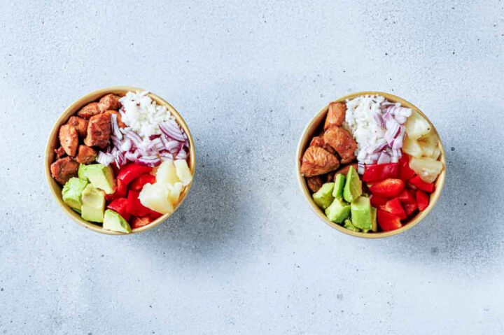Overhead view of two fully assembled Hawaiian Chicken Bowls placed side-by-side. Each bowl contains white rice, cooked chicken, diced avocado, red bell pepper, cubed pineapple, and sliced red onion.