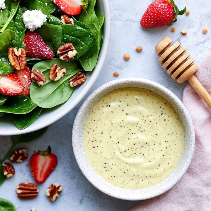 A bowl of creamy honey mustard dressing with black pepper, served next to a spinach salad topped with sliced strawberries, pecans, and feta cheese.
