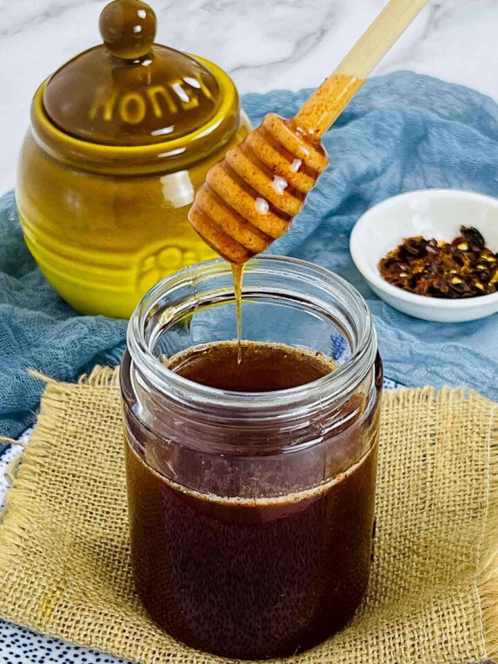 A wooden honey dipper lifts dark amber hot honey from a glass jar, with a ceramic honey pot and a bowl of chili flakes in the background.