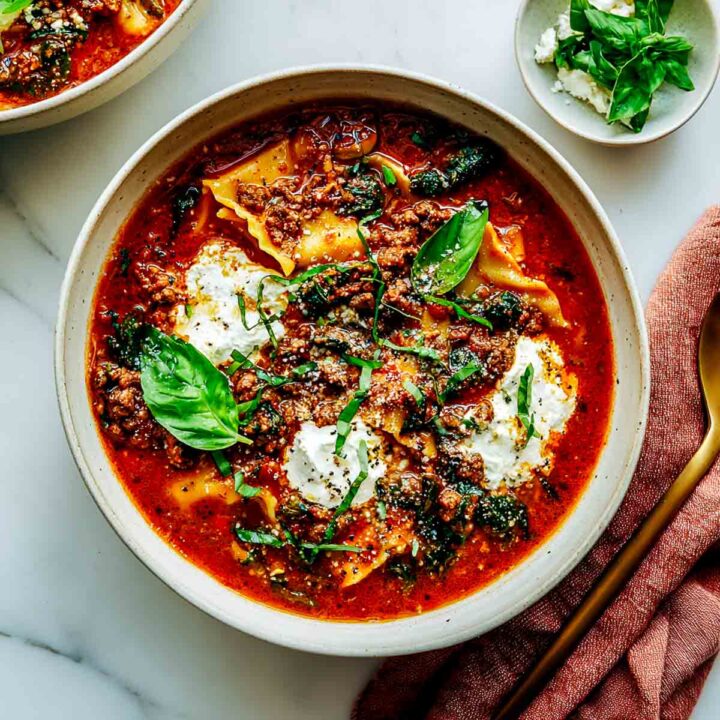 A bowl of rich lasagna soup with a tomato and meat or plant-based sauce, pasta sheets, dollops of ricotta cheese, and fresh basil leaves.