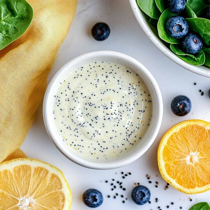 A small bowl of creamy lemon poppy seed Greek yogurt dressing, surrounded by blueberries, a sliced lemon, and a sliced orange.
