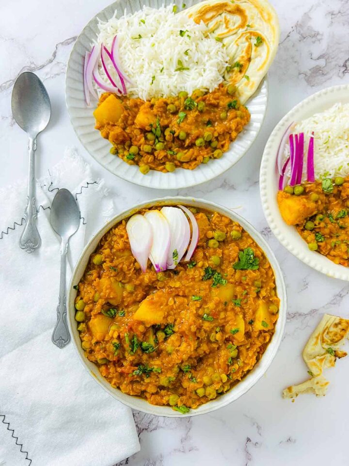 Overhead view of a bowl of lentil, pea, and potato curry (Aloo Matar Dal) garnished with sliced red onion, with two plates of the curry, rice, and naan bread on the side.