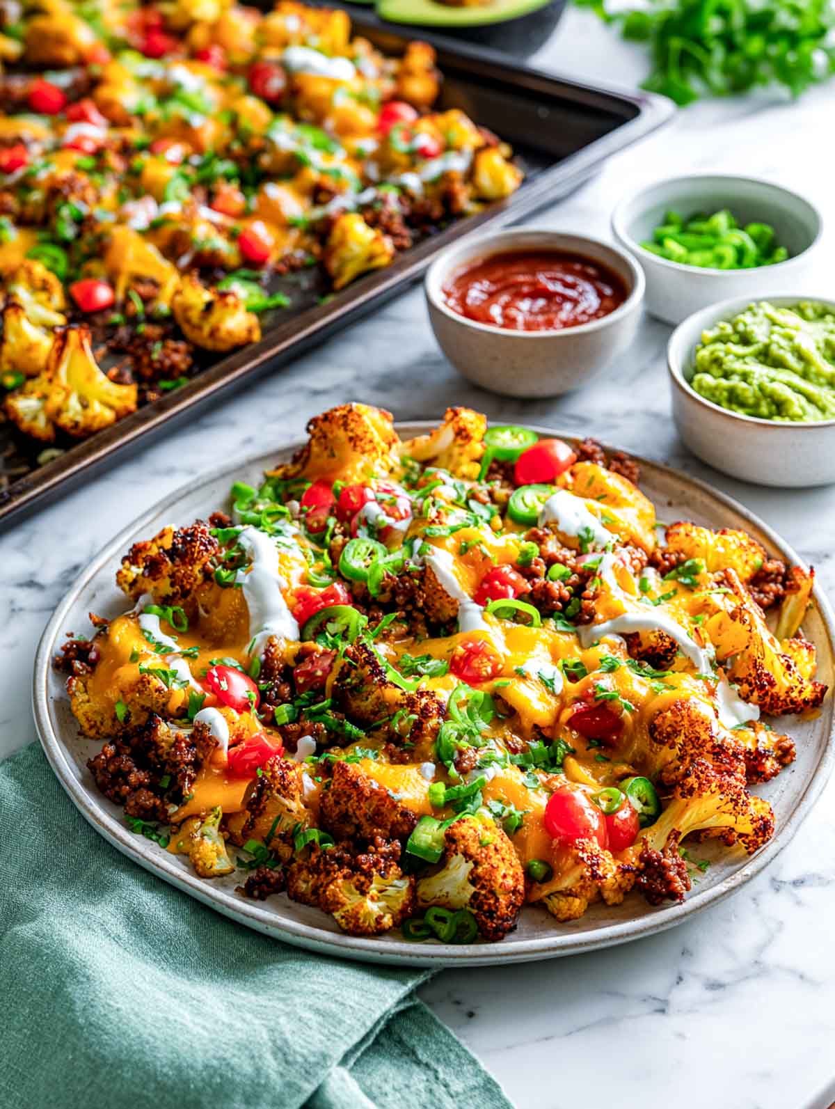 Plated loaded cauliflower nachos topped with melted cheese, ground turkey, tomatoes, jalapeños, and sour cream on a marble surface with guacamole and salsa bowls beside.