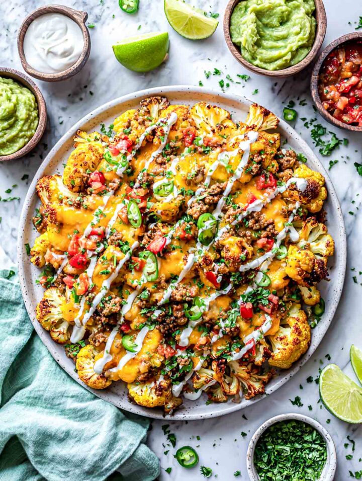 Top-down view of plated high protein loaded cauliflower nachos with melted cheese, seasoned turkey, and fresh toppings, surrounded by bowls of guacamole, salsa, and lime wedges on marble surface.