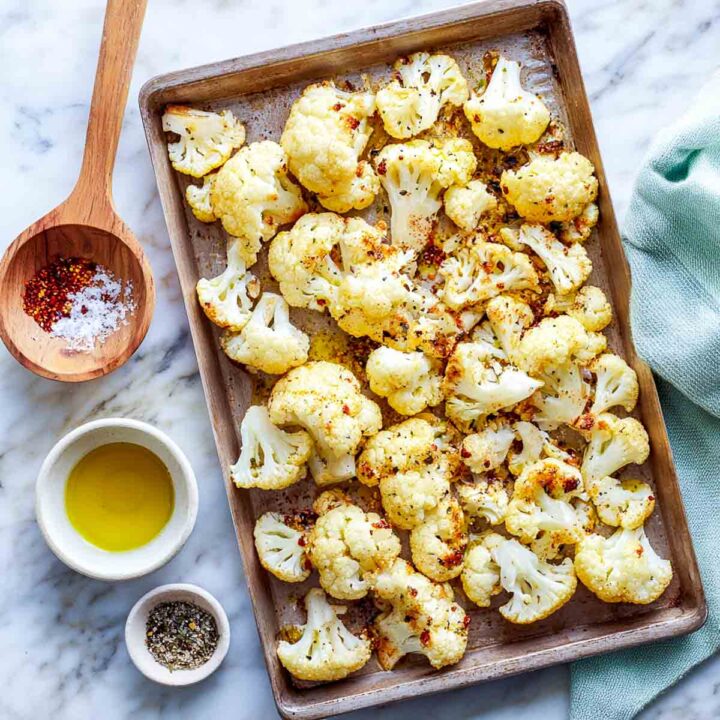 Baking tray of cauliflower florets seasoned with olive oil, salt, pepper, and paprika before roasting, on marble background with a light green napkin.