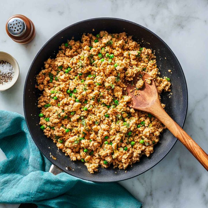 Top view of ground turkey mixture with peas cooking in skillet on marble surface with teal linen and wooden spoon.