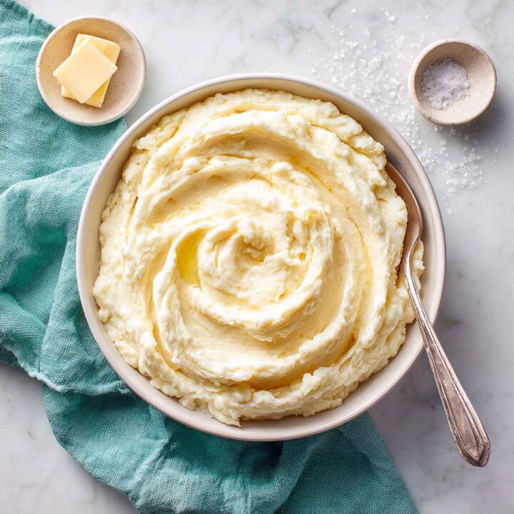 Bowl of creamy cauliflower mash with melted butter swirl on marble surface and teal linen beside it.