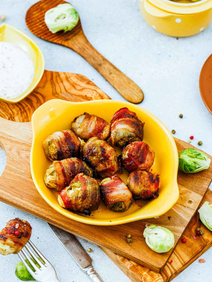 A small yellow oval dish holding several oven-roasted Brussels sprouts wrapped in bacon, glazed with maple syrup. The dish sits on a wooden cutting board, with fresh Brussels sprout halves, scattered peppercorns, a small bowl of white dipping sauce, and a wooden spoon visible in the rustic, light-colored background.