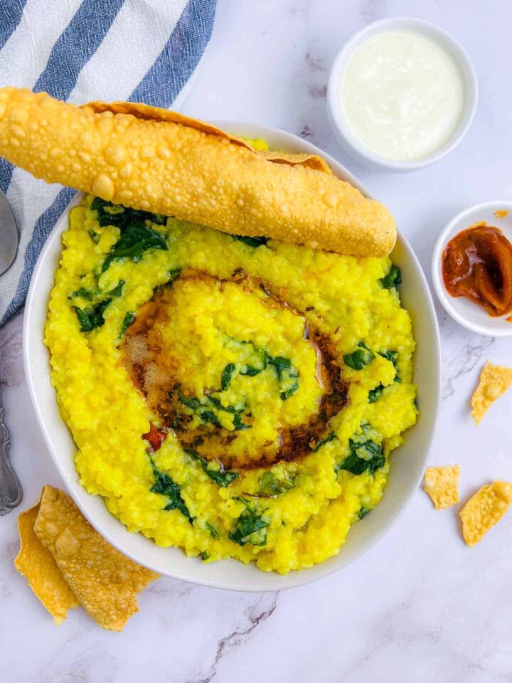 Yellow palak khichdi (rice and lentil porridge with spinach) in a white bowl, topped with a drizzle of oil or ghee and served with a fried papadum, yogurt, and a small dish of pickle or chutney.