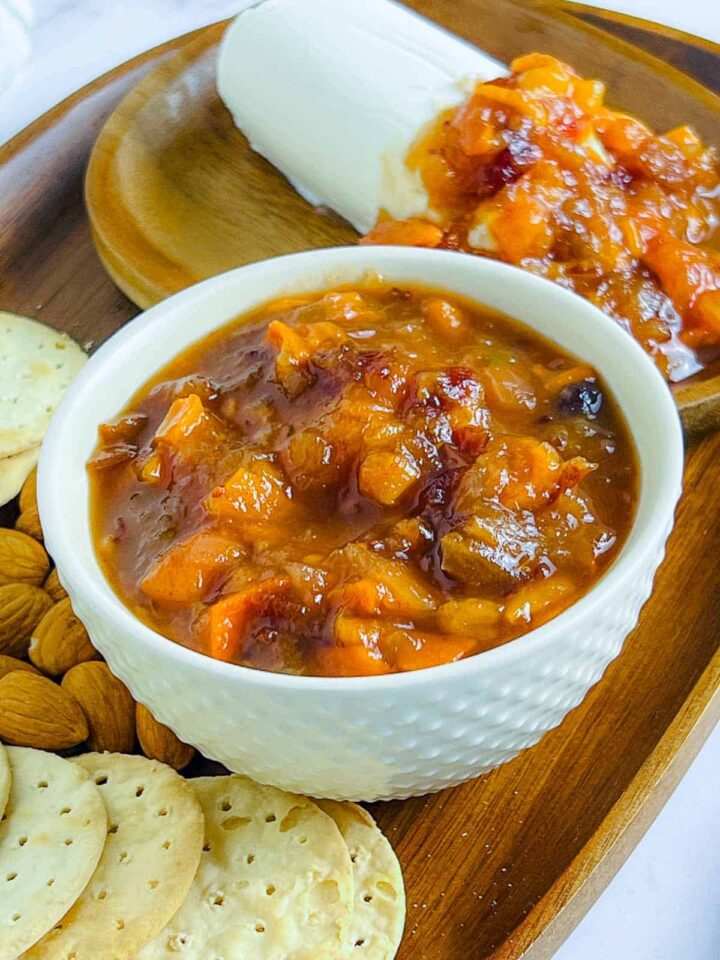 A small white bowl of chunky orange persimmon and cranberry chutney, served on a wooden board with goat cheese, almonds, and crackers.
