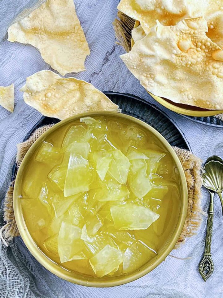 A bowl of translucent yellow 'plastic' chutney (papaya or bottle gourd) with syrupy liquid, served with large, fried papadums.