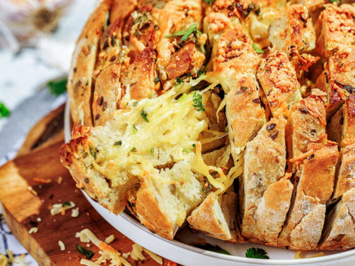 Close-up shot of a round loaf of pull-apart garlic bread (or blooming bread) that has been deeply scored and filled with melted, stringy cheese and herbs. The golden, crispy bread sits on a white plate over a wooden cutting board.
