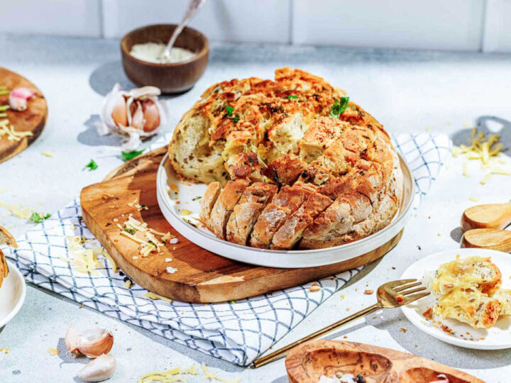 A finished, crusty pull-apart garlic bread loaf, stuffed with melted cheese and herbs, is served on a white plate atop a wooden cutting board and a checkered cloth. Various elements of the preparation, including garlic cloves, shredded cheese, a bowl of Parmesan, and a golden fork, are scattered around the bread on a bright, speckled countertop.