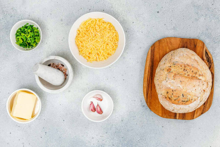 Overhead flat lay of ingredients for pull-apart garlic bread laid out on a gray countertop: a round loaf of bread sits on a wooden board next to bowls containing chopped herbs, shredded yellow cheese, a mortar and pestle with salt, two cloves of garlic, and a stick of butter.