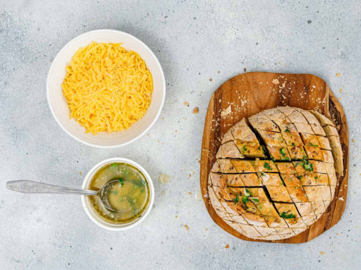 Overhead shot showing the first step in assembling pull-apart garlic bread: a round loaf of scored bread is brushed with a seasoned butter/herb mixture on a wooden board. Next to it are a bowl of shredded cheese and a small bowl of the garlic butter mixture with a spoon.