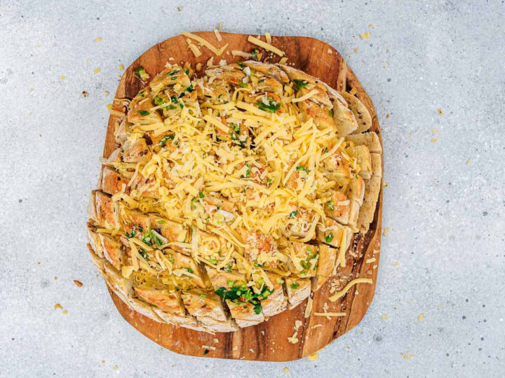 Overhead shot showing a round loaf of scored bread already brushed with garlic butter, now heavily topped and stuffed with shredded cheddar cheese and chopped herbs, ready for baking.