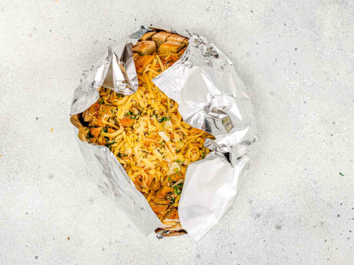 Overhead shot of a round loaf of pull-apart garlic bread, generously stuffed with cheese and herbs, wrapped loosely in aluminum foil, revealing the top section of the filling, ready for baking.