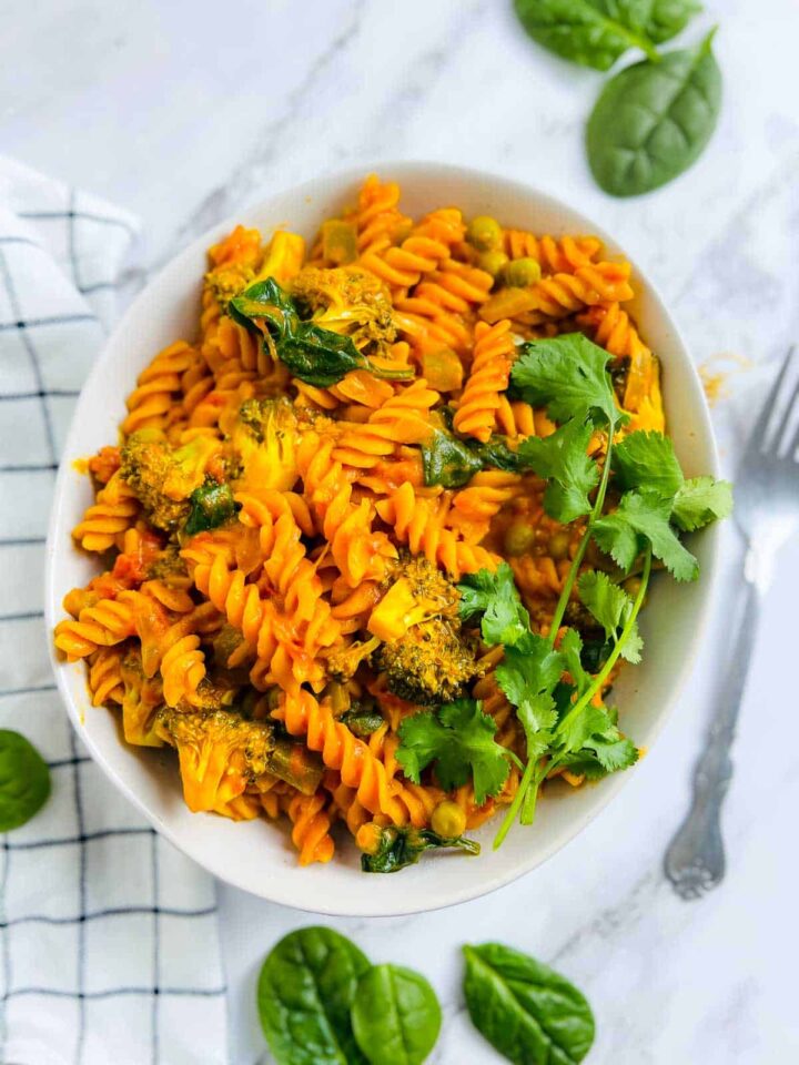 Pasta with red curry sauce, broccoli, and spinach, served in a white bowl and garnished with fresh cilantro.