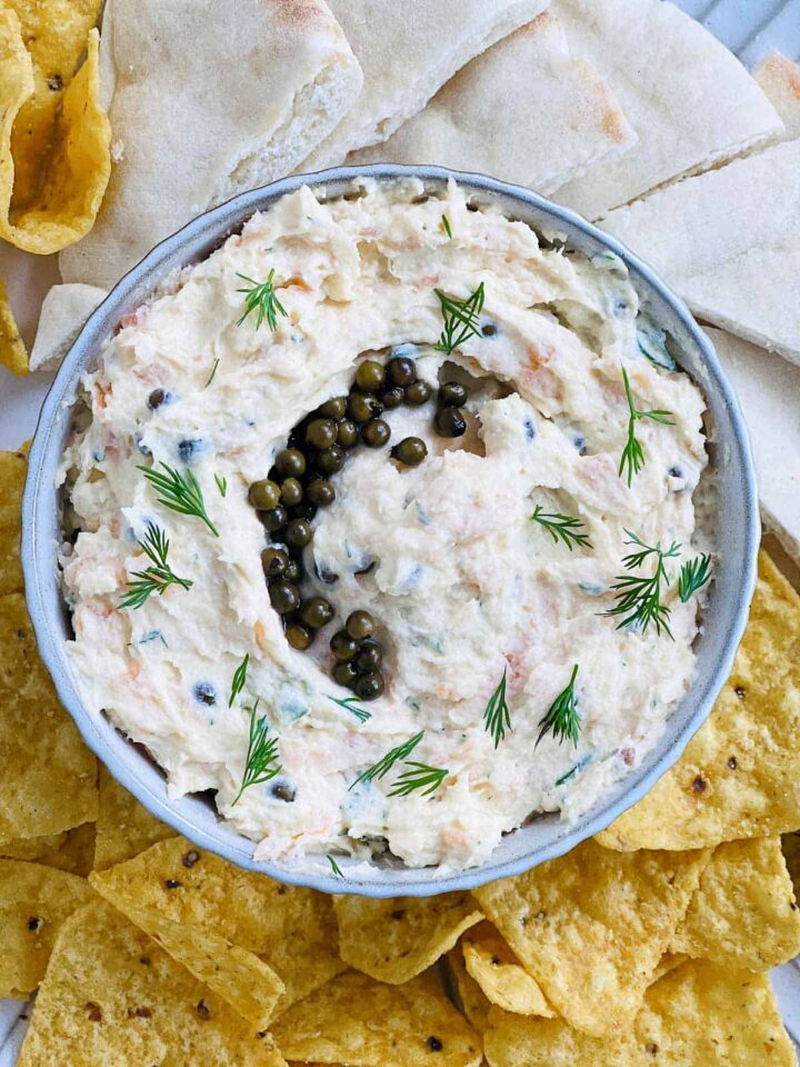 An overhead close-up shot of a bowl of creamy salmon and egg dip garnished with fresh dill sprigs and a ring of caper berries in the center. The dip is surrounded by corn tortilla chips and wedges of pita bread.