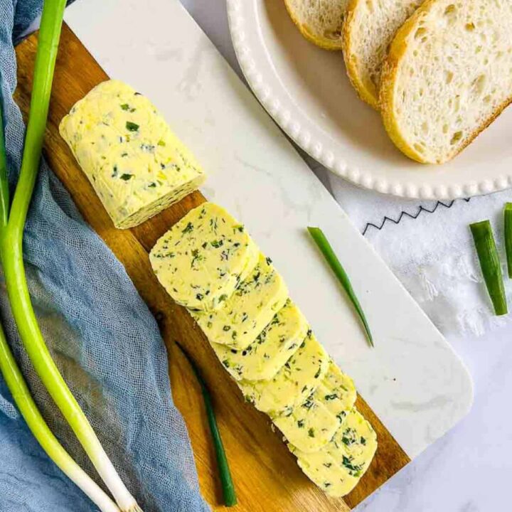 A log of bright yellow scallion or chive compound butter, partially sliced, served on a marble and wood board next to slices of bread.
