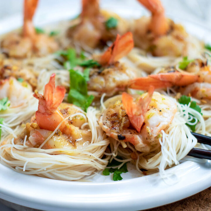 A close-up of a white plate holding seven cooked, peeled shrimp with tails attached, each resting in a small nest of fine vermicelli noodles. The dish is seasoned with a garlic-herb sauce and garnished with fresh parsley, with chopsticks and a small dipping bowl visible nearby.