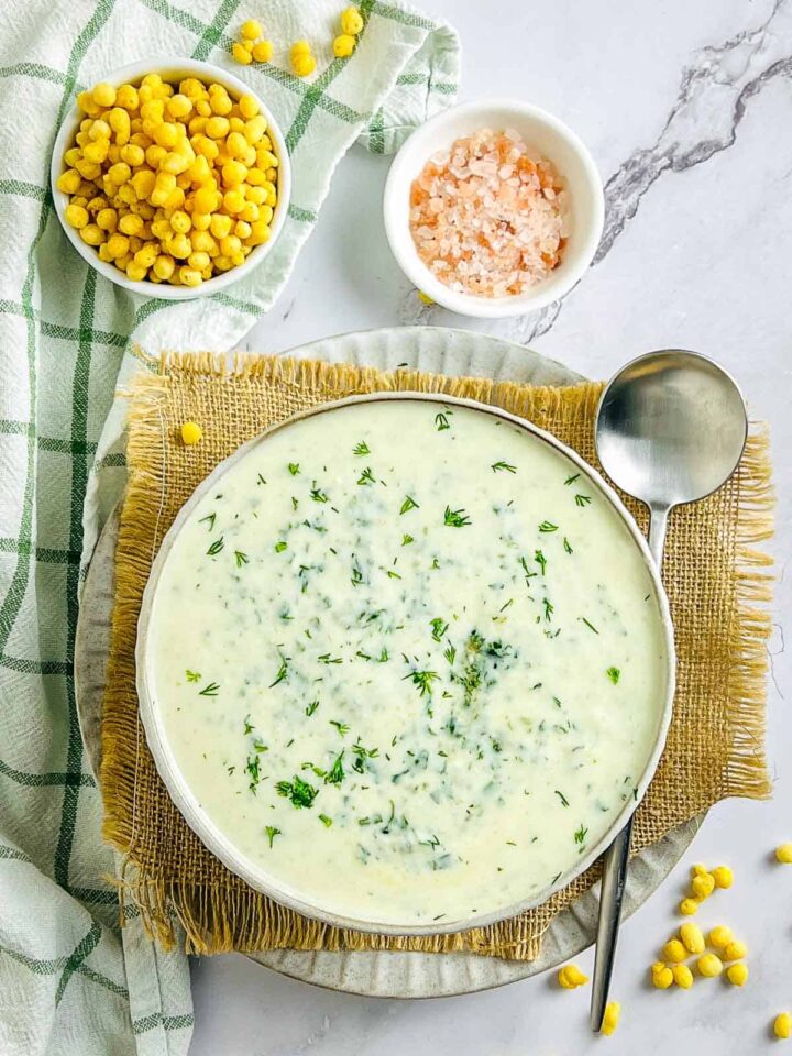 An overhead shot of a bowl of creamy white spinach and dill raita garnished with fresh dill, served on a burlap mat with a metal ladle. Two small bowls containing crunchy yellow snacks (likely boondi or seasoned corn) and pink Himalayan salt are placed nearby.