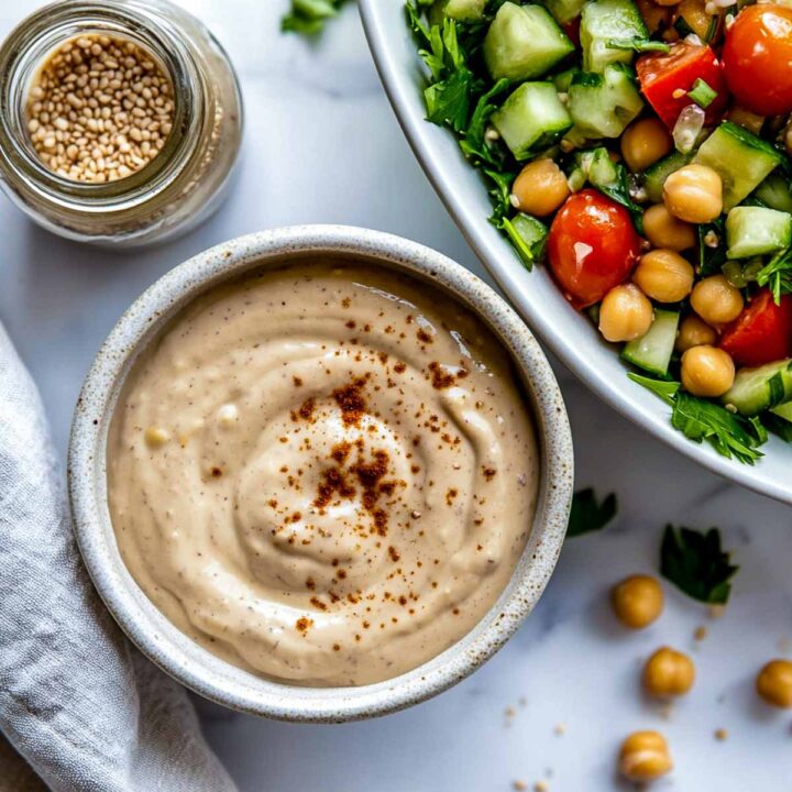 A bowl of creamy tahini and Greek yogurt dressing, topped with paprika or chili powder, served next to a chickpea and vegetable salad.