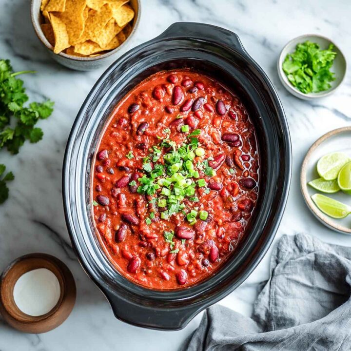 Overhead shot of three-bean chili in a black slow cooker, garnished with chopped green onions, and served with sides of cilantro, lime wedges, and tortilla chips.