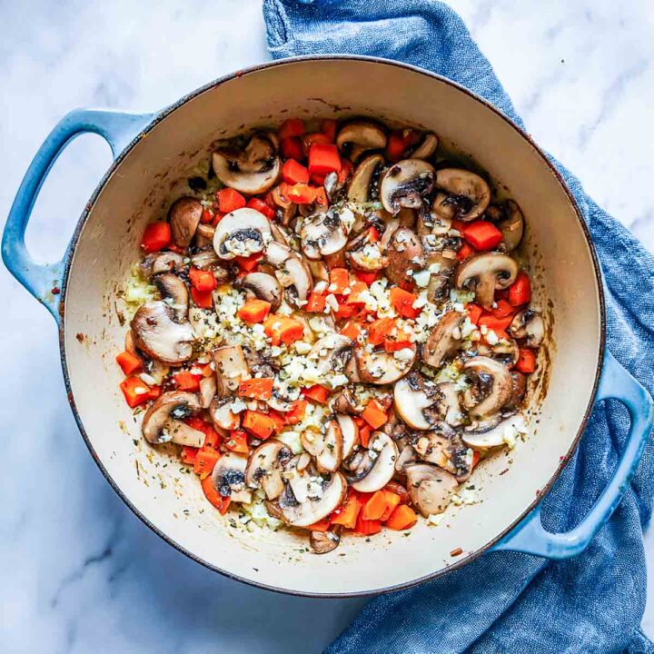 Mushrooms, carrots, and garlic sautéing in Dutch oven on marble surface.