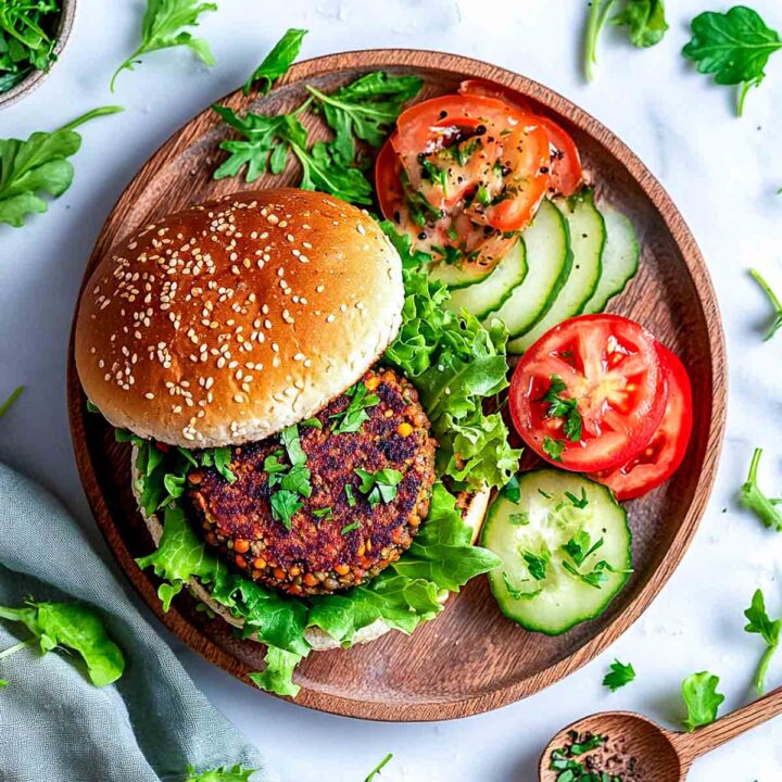 Vegan bean and lentil burger on a sesame bun with lettuce, served on a wooden plate with sliced tomatoes and cucumbers.