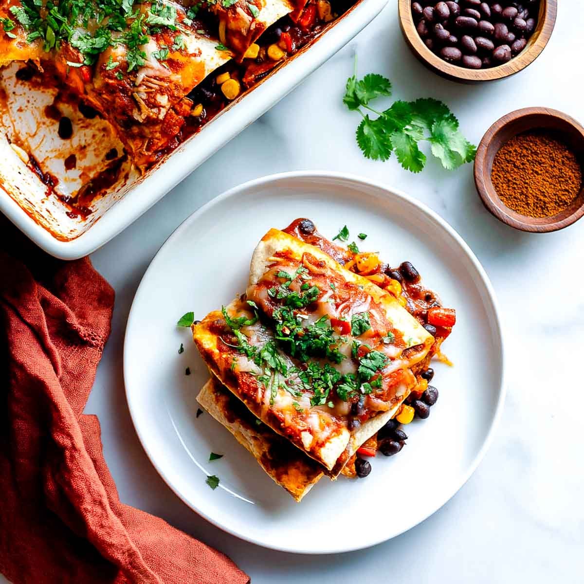 Two vegan enchiladas topped with red sauce, melted cheese, and fresh cilantro, served alongside a white baking dish.