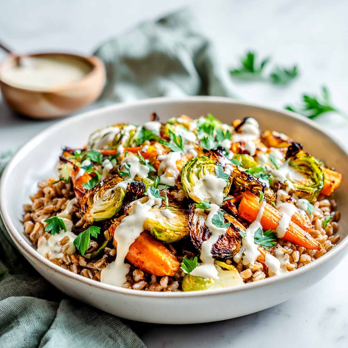 Warm farro bowl topped with roasted Brussels sprouts and carrots, drizzled with a creamy dressing and garnished with fresh parsley.