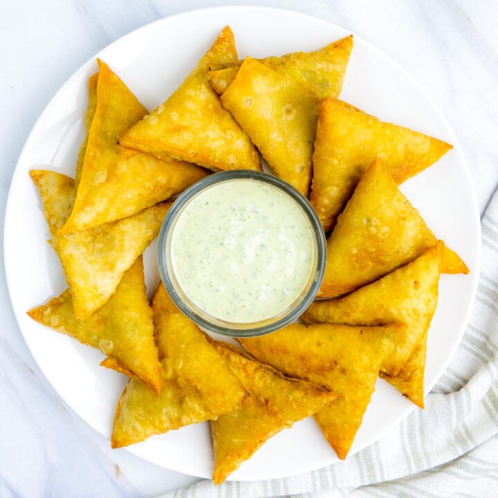 An overhead view of a circular white plate arranged with over a dozen golden-brown, triangular aloo samosas circling a small glass bowl of light green mint or cilantro chutney in the center. The plate sits on a white marble surface next to a striped linen napkin.